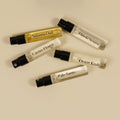 Five clear glass perfume sample vials with black spray caps arranged on a warm beige background. The vials contain different colored liquids and are labeled with scents: Satsuma Oud, Hinoki Sesame, Caesars Flower, Desert Kush, and Palo Santo.