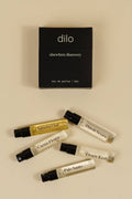 Five small perfume sample vials with black spray caps are arranged on a warm beige background below a black rectangular box labeled 'dilo elsewhere discovery eau de parfum / 5 ml'. The vials contain clear, yellow, and white liquids, with some labels partially visible including 'Sakura Oud', 'Citrus Flower', 'Desert Kush', and 'Pale Samso'.
