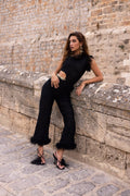 A woman models a black sleeveless jumpsuit with feathered trim and wide flared legs, leaning against a stone wall in an outdoor setting.
