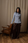 A woman stands in a room with beige curtains and a wooden floor, wearing a light blue and white vertically textured long-sleeve button-up shirt and dark brown wide-leg trousers with white side stripes. She is standing next to a brown leather and wood armchair.