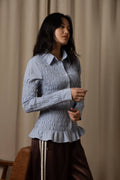 A woman wearing a light blue and white vertically striped, smocked long-sleeve blouse with a ruffled hem, paired with dark brown pants, standing in front of a beige curtain backdrop.