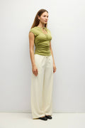 A woman modeling a fitted olive green short-sleeve top with a keyhole cutout and ruched sides, paired with wide-leg cream pants and black flats, standing against a plain light gray studio background.