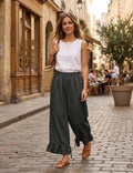A woman walking on a cobblestone street wearing a white sleeveless top, dark gray wide-leg linen pants with ruffled hems, and tan leather sandals, carrying a woven straw bag.