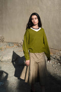 A woman stands outdoors against a textured stucco wall wearing an olive green long-sleeve sweater with a white V-neck trim and a beige midi skirt.