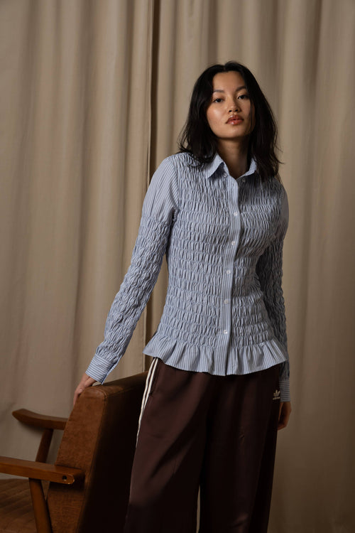 A woman stands indoors wearing a light blue-gray smocked long-sleeve button-up shirt with a ruffled hem and dark brown wide-leg pants. She is posed in front of beige curtains, leaning against a wooden chair.