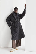 A model wearing a black quilted puffer wrap coat with a folded front collar and a self-belt with loops on the side. The coat has hidden pockets and is oversized for comfort.
