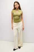 A woman modeling a sage green ruched short-sleeve top with a tie-neck detail, paired with cream wide-leg drawstring pants and black shoes, against a plain light gray studio background.