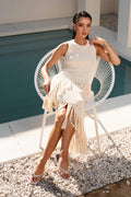 Woman in a white sleeveless top and fringe skirt sitting by a pool.