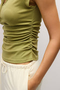 Close-up of a person wearing an olive green ruched tank top and off-white drawstring pants against a light gray background.