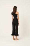 A woman viewed from the back wearing a black one-shoulder lace jumpsuit with feather trim at the neckline and hem, paired with gold high-heeled sandals, against a plain off-white studio background.