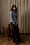 A woman posing in a studio setting wearing a light blue textured long-sleeve button-up shirt and dark brown wide-leg pants, standing next to a wooden chair against a beige curtain backdrop.
