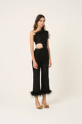 A model stands against a plain off-white studio background wearing a black one-shoulder lace jumpsuit with feather trim at the neckline and hem, a gold waist detail, and gold heeled sandals.