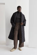 A model wearing a black quilted puffer wrap coat with a folded front collar and a self-belt with loops on the side. The coat has hidden pockets and is oversized for comfort.