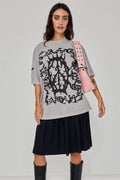 A model stands against a light gray studio background wearing an oversized light gray graphic T-shirt with a large black skull-and-scroll design, a dark pleated skirt, and black knee-high boots. She carries a pink shoulder bag.