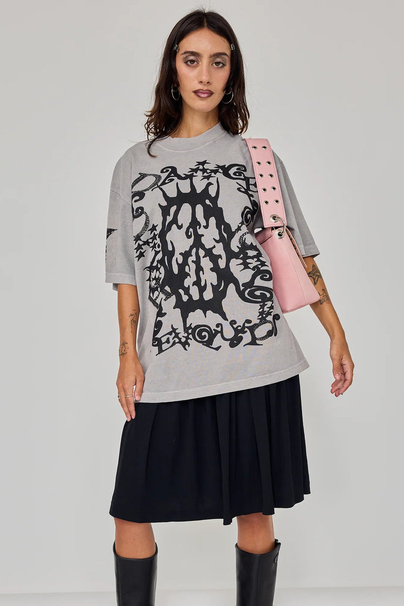 A model stands against a light gray studio background wearing an oversized light gray graphic T-shirt with a large black skull-and-scroll design, a dark pleated skirt, and black knee-high boots. She carries a pink shoulder bag.