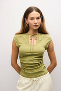 A woman modeling a fitted olive green short-sleeve top with a keyhole tie neckline, paired with cream drawstring pants, against a plain light gray studio background.