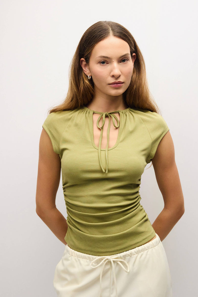 A woman modeling a fitted olive green short-sleeve top with a keyhole tie neckline, paired with cream drawstring pants, against a plain light gray studio background.