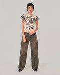 A model stands against a light gray studio background wearing a beige short-sleeve top with black floral embroidery and black-and-white gingham check accents on the sleeves, paired with dark gray wide-leg jeans featuring silver stud detailing along the seams.