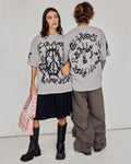 Two models wearing oversized light gray graphic T-shirts with black gothic-style prints. One model shows the front of the shirt with a black skirt and black boots, holding a pink bag.