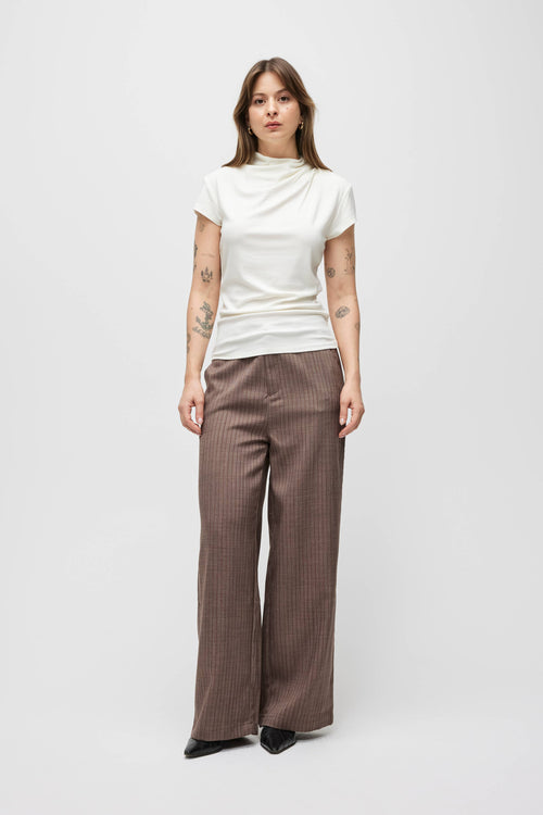 A model stands against a light gray studio background wearing a white mock-neck short-sleeve top and brown pinstriped wide-leg trousers.