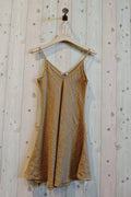 Gold slip dress hanging on a wooden hanger against a wooden wall.