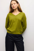 A woman wearing a loose olive green V-neck knit sweater and dark navy pants, standing against a plain light gray wall.
