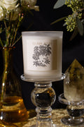Candle with decorative label on a glass holder next to a vase of flowers and a crystal.