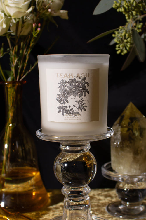 Candle with decorative label on a glass holder next to a vase of flowers and a crystal.