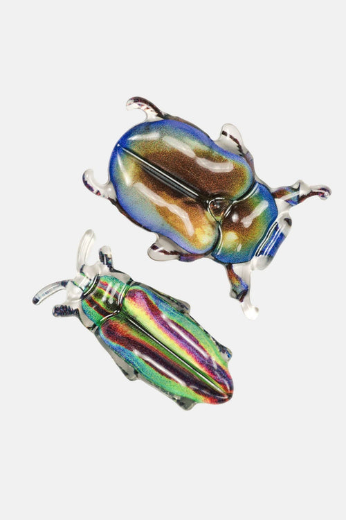 Two iridescent beetle-shaped brooches with metallic rainbow reflections on a plain light gray background.