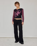 A model stands in a studio wearing a black long-sleeve crop top with pink and black striped sleeves, a pink skull graphic, and black pinstriped wide-leg pants.