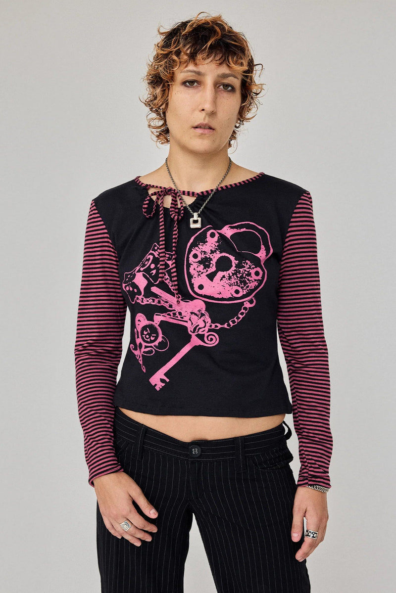 A model wearing a black long-sleeve crop top with pink and black horizontal striped sleeves, featuring a large pink graphic of a skull and key on the front, paired with black pinstriped pants.